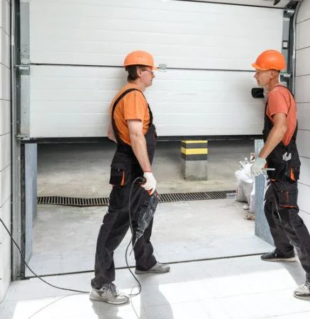 Two workers in orange hard hats installing a white garage door.