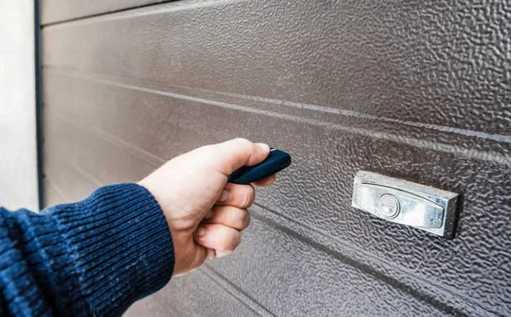 Hand holding a garage door opener remote near a brown garage door.