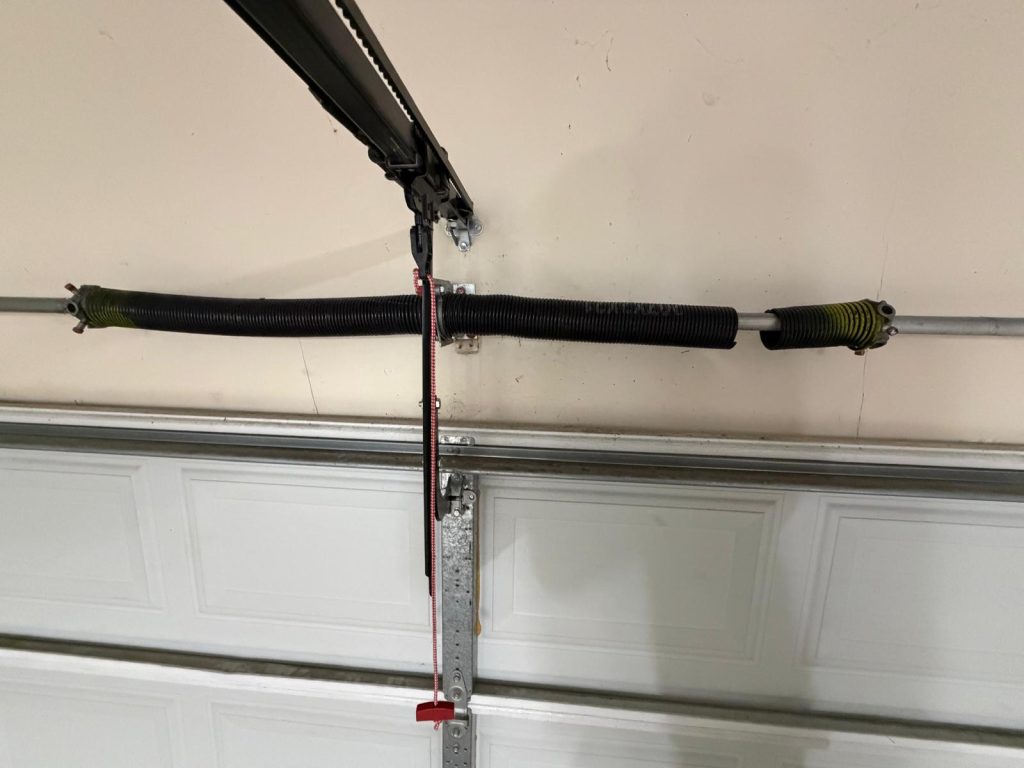 Broken garage door spring and opener mechanism above a white garage door.