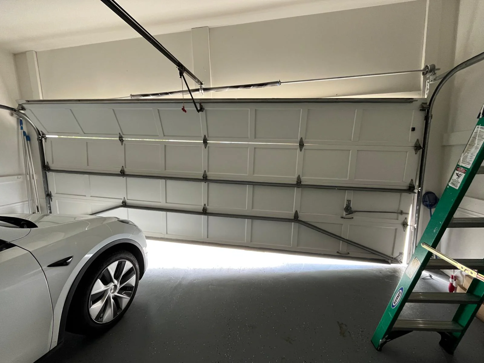 White garage door partially open with a white car and green ladder