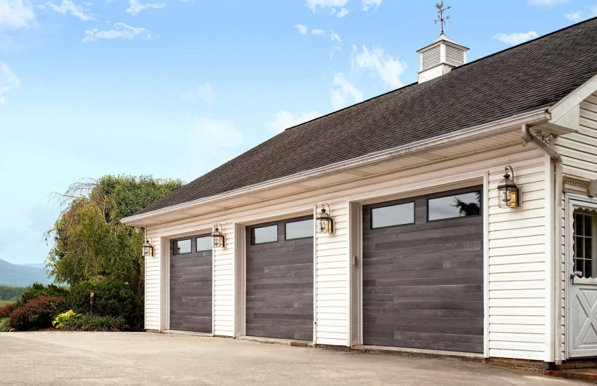 Three dark gray garage doors on a white house under a blue sky
